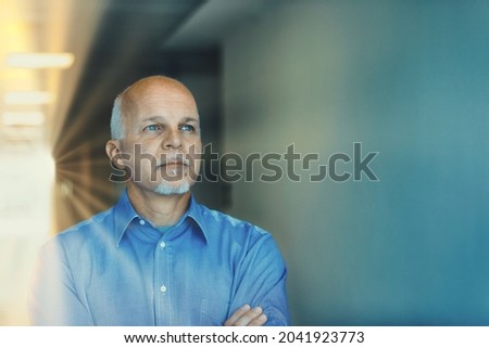 Conceptual image of senior man with forethought, creativity and vision standing with folded arms staring ahead deep in thought backlit by rays from a sunburst in an office building