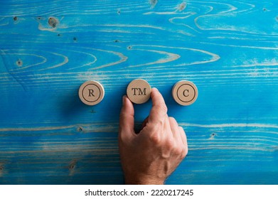 Conceptual image of trademark and copyrights - male hand placing three wooden cut circles with circled letter signs R, C and TM on them over textured blue wooden background.