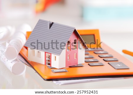 Conceptual Miniature Model Home on Top of an Orange Calculator Device Placed on White Table with Blueprint.