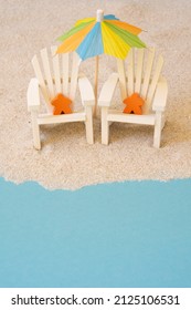 A Conceptual Photograph Of Miniature Wooden People Sitting On Adirondack Chairs In The Sand And Under An Umbrella At The Beach.