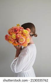 Conceptual Portrait Of Young Woman Wearing White Cotton Dress Covering Her Face With Lush Ranunculus. Female Hiding Behind Beautiful Bouquet Of Bright Flowers. Close Up, Copy Space, White Background