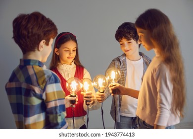 Conceptual Shot Of Happy Clever Kids Standing In Circle Holding Glowing Light Bulbs. Team Of Young School Children Join Shining Edison Lightbulbs As Metaphor For Sharing And Developing Creative Ideas