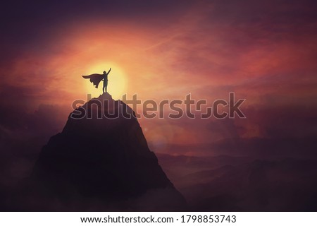 Conceptual sunset scene, superhero with cape standing brave on top of a mountain looks determined at horizon raising one hand up as a winning leader. Hero power and motivation, overcoming obstacles.