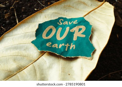 Conceptual view of a dark green torn paper with handwritten Save OUR earth message resting on a dry leaf. Perfect for environmental protection and eco activism banners