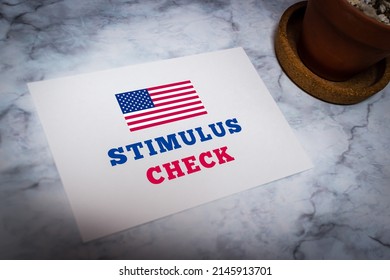 Conceptual White Message Card Showing The Message “Stimulus Check” With American Flag On White Marble Table With Cactus.