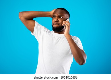 Concerned African American Man Talking On Phone Touching Head In Stress Having Problems Posing Standing Over Blue Background In Studio. Unpleasant Mobile Call Concept