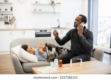 Concerned African dad checks temperature of sick young boy while calling doctor for advice. Father and son are in cozy living room with medicine on table. - Powered by Shutterstock