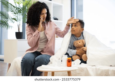 Concerned African mother checks temperature of sick young boy using thermometer while holding phone to consult doctor. Boy, wrapped in blanket, clutches teddy bear for comfort. - Powered by Shutterstock