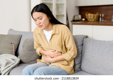 Concerned Asian Woman Suffering From Stomach-ache Sitting On Sofa. Female Holding Tummy, Undergoing Belly Pain And Discomfort, Suffer From Menstruation
