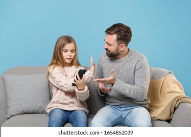 Concerned Bearded Man In Knitted Sweater With Child Baby Girl. Father Little Daughter Isolated On Pastel Blue Background. Love Family Parenthood Childhood Concept. Sitting On Couch, Hold Mobile Phone