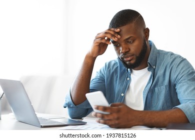 Concerned Black Freelancer Guy Looking At Smartphone While Working At Home Office