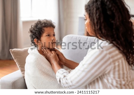 Concerned black mother gently touching her son's throat, checking for signs of illness while he sits wrapped in blanket on couch, mom caring about her sick preteen kid at home, closeup