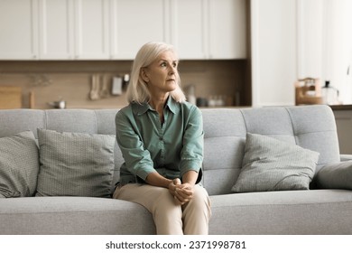 Concerned blonde senior woman sitting on couch in modern home interior, looking at window away, thinking on problems, bad anxious news, suffering from stress, feeling nervous - Powered by Shutterstock