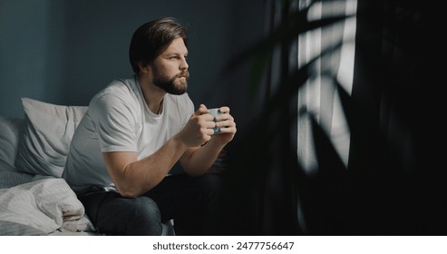 Concerned Caucasian man sittind on the bed near window with cup of tea. Young male drinking hot coffee looking outside at landscape. - Powered by Shutterstock