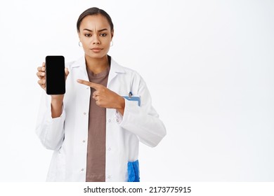 Concerned Doctor Pointing Finger At Mobile Phone Screen, Frowning And Looking Serious, Standing Over White Background. Online Medical Clinic App