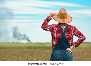 Concerned Female Farmer In Corn Field Looking At Black Smoke On Horizon, Concept Of Insurance In Agriculture And Farming