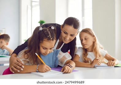 Concerned female teacher helping little schoolgirls with lesson in primary school classroom. Smiling woman teaches children spelling. Children write in notebooks. Concept of junior education. - Powered by Shutterstock