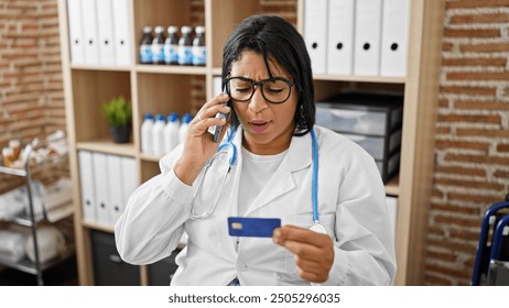 A concerned hispanic woman doctor examines a credit card while talking on the phone in a clinic office. - Powered by Shutterstock