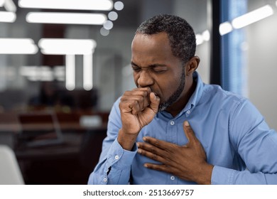 Concerned man coughing at work with hand on chest, indicating cold. Seated in modern office setting wearing blue shirt, appears uncomfortable, reflecting health concern or stress in workplace. - Powered by Shutterstock