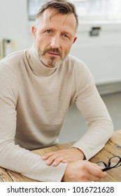 Concerned man with an earnest expression staring intently at the camera as he sits at a table holding his glasses