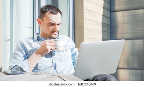 Concerned Man Having A Facetime Video Call Talking To His Colleagues. Unhappy Sad Businessman Having A Video Conference Outside On A Sunny Day. Self Isolating At Home On Balcony Drinking Coffee. 