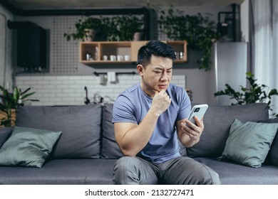 Concerned Man Reading News Online Looking At Phone Screen, Asian Sitting On Sofa At Home, Serious And Sad