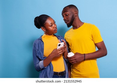 Concerned Man Spying On Girlfriend Sms On Mobile Phone. Young Woman Holding Smartphone And Browsing Internet While Her Boyfriend Looks At Notification On Screen In Studio, Expecting Betrayal.