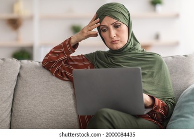 Concerned Middle-Aged Muslim Lady Using Laptop Reading Negative News Online Sitting On Sofa At Home. Concerned Middle-Eastern Woman Having Problem At Work Browsing Internet On Computer