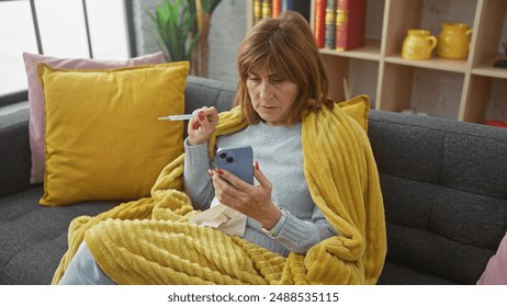 A concerned middle-aged woman checks her temperature with a thermometer while using a smartphone on the couch at home. - Powered by Shutterstock