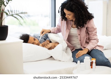 Concerned mother of African descent checks temperature of her son, young African American boy, using thermometer. Mother sits next to the bed where child rests, showing care and comfort. - Powered by Shutterstock