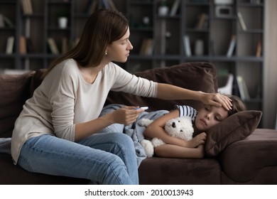 Concerned mother checking high body temperature of daughter, touching head, forehead, holding thermometer. Sad sick girl lying on sofa, suffering from influenza, fever. Children healthcare concept