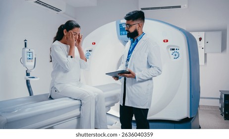 Concerned patient pointing at screen while asking question. Calm bearded doctor in medical coat holding tablet and responding while standing beside modern scanning equipment in hospital room.