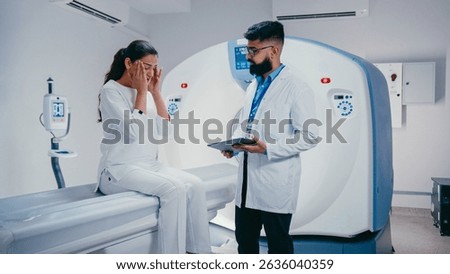 Concerned patient pointing at screen while asking question. Calm bearded doctor in medical coat holding tablet and responding while standing beside modern scanning equipment in hospital room.