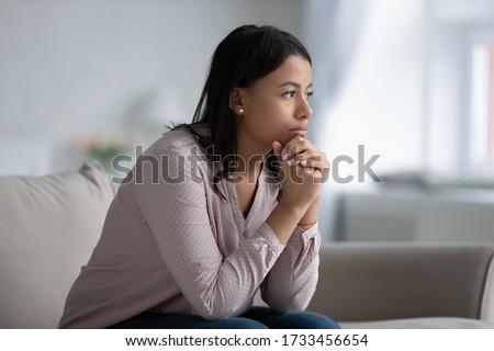 Concerned sad African woman sit on sofa in living room thinking of problem looks in distance feels depressed due loneliness, inner emptiness, life troubles, break up or divorce, marriage split concept