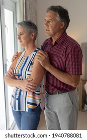 Concerned Senior Caucasian Couple Standing Looking Out Of Window Embracing. Staying At Home In Isolation During Quarantine Lockdown.