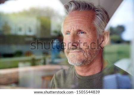 Concerned Senior Man Standing And Looking Out Of Kitchen Door Viewed Through Window