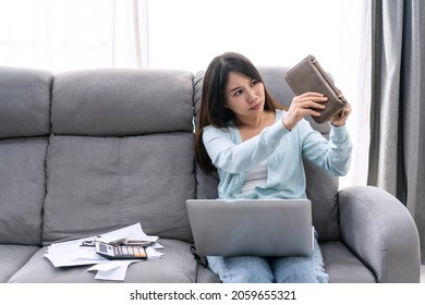 Concerned Stressed Young Asian Woman Sitting On Sofa At Home Looking Her Empty Purse With Paper Letters From Bank Informing About Debt Bankruptcy. Financial, Unprofitable, Debt Concept.