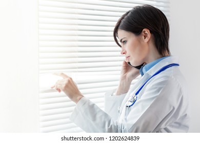 Concerned Thoughtful Woman Doctor Standing In Front Of A Window With Louver Blinds Staring Down Talking On A Mobile Phone With Copy Space