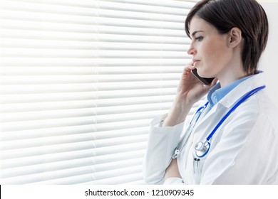 Concerned Thoughtful Woman Doctor Standing In Front Of A Window With Louver Blinds Staring Down Talking On A Mobile Phone With Copy Space