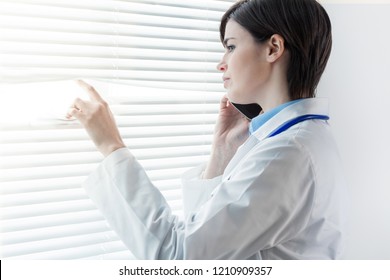 Concerned Thoughtful Woman Doctor Standing In Front Of A Window With Louver Blinds Staring Down Talking On A Mobile Phone With Copy Space