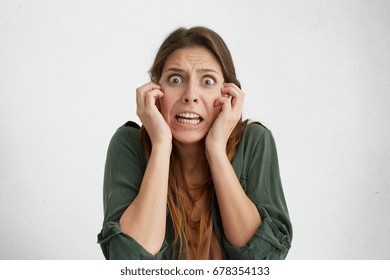 Concerned Woman With Bugged Dark Eyes Keeping Hands On Cheeks Looking Desperately Being Scared Of Something Horrorful. Shocked Female Looking Terrified. Facial Expression And Emotions Concept