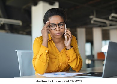 Concerned young black businesswoman talking on cellphone and touching head in stress, worried female entrepreneur suffering problems with work, having unpleasant mobile call