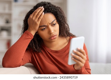 Concerned young black woman looking at smartphone screen and touching head, upset african american female sitting on couch with troubled expression, suffering stress or got bad news on phone - Powered by Shutterstock