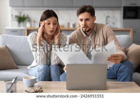 Concerned young spouses intensely scrutinize papers while sitting on couch with laptop open, indicating they are dealing with serious matters