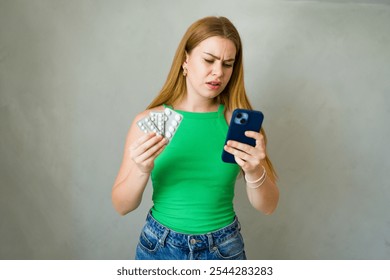 Concerned young woman holding blister packs of pills and reading instructions on her phone with a worried expression - Powered by Shutterstock