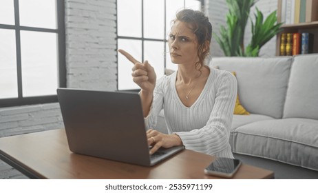 A concerned young woman points at a laptop screen while sitting in a modern living room, indicating an issue or question. - Powered by Shutterstock