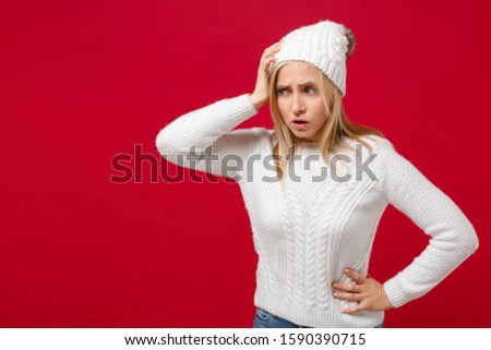 Concerned young woman in white sweater, hat isolated on red wall background, studio portrait. Healthy fashion lifestyle cold season concept. Mock up copy space. Putting hand on head, looking aside