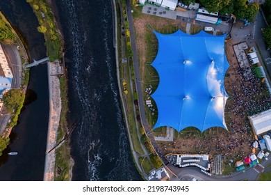 Concert Of Beer Festival Area With Big Blue Tent And Service Areas By A River. Big Crowd Enjoy All Amenities. Aerial Top Down View. Music Event Or Circus In Town. Entertainment Industry And Business.