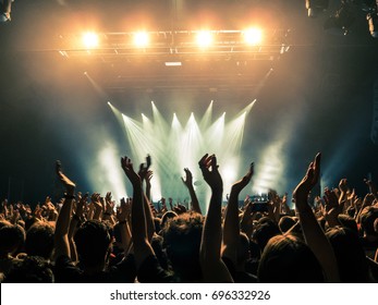 Concert crowd attending a concert, people silhouettes are visible, backlit by stage lights. Raised hands and smart phones are visible here and there.