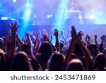 Concertgoers raise hands in rhythm at outdoor music festival, colorful stage lights illuminate excited audience. Fans enjoy live performance, nightlife energy, summer event brings people together.